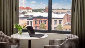 A hotel room with a laptop on a round table, checkered chairs, a vase with flowers, and a view of buildings and a church through large windows.