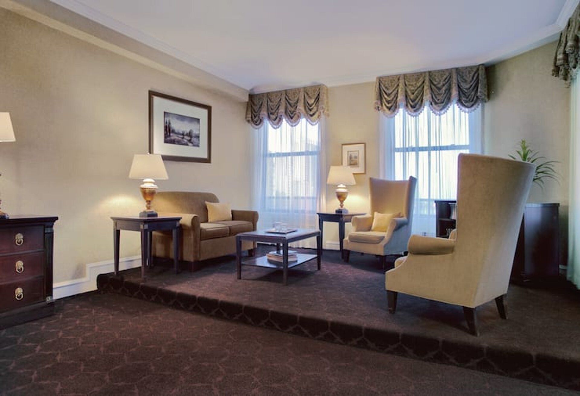 A living room with a sofa, armchair, coffee table, and lamps. There are two windows with curtains, a picture on the wall, and a plant.