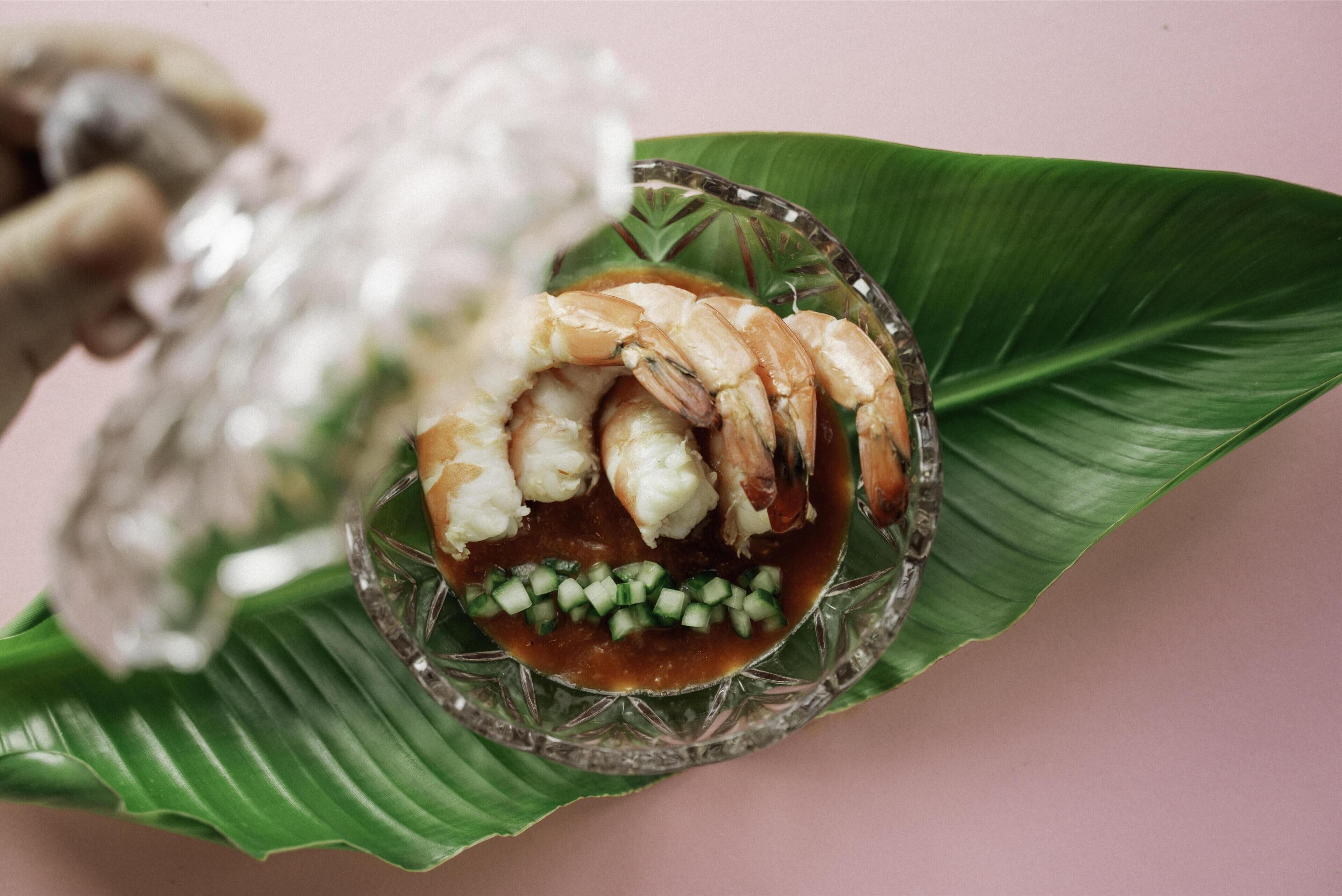 A glass bowl with shrimp on a leaf, accompanied by a glass container of ice.