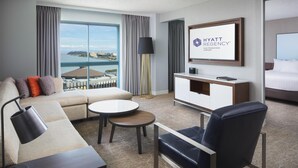 A modern hotel room with a sofa, armchair, dining table, and a view of the city.