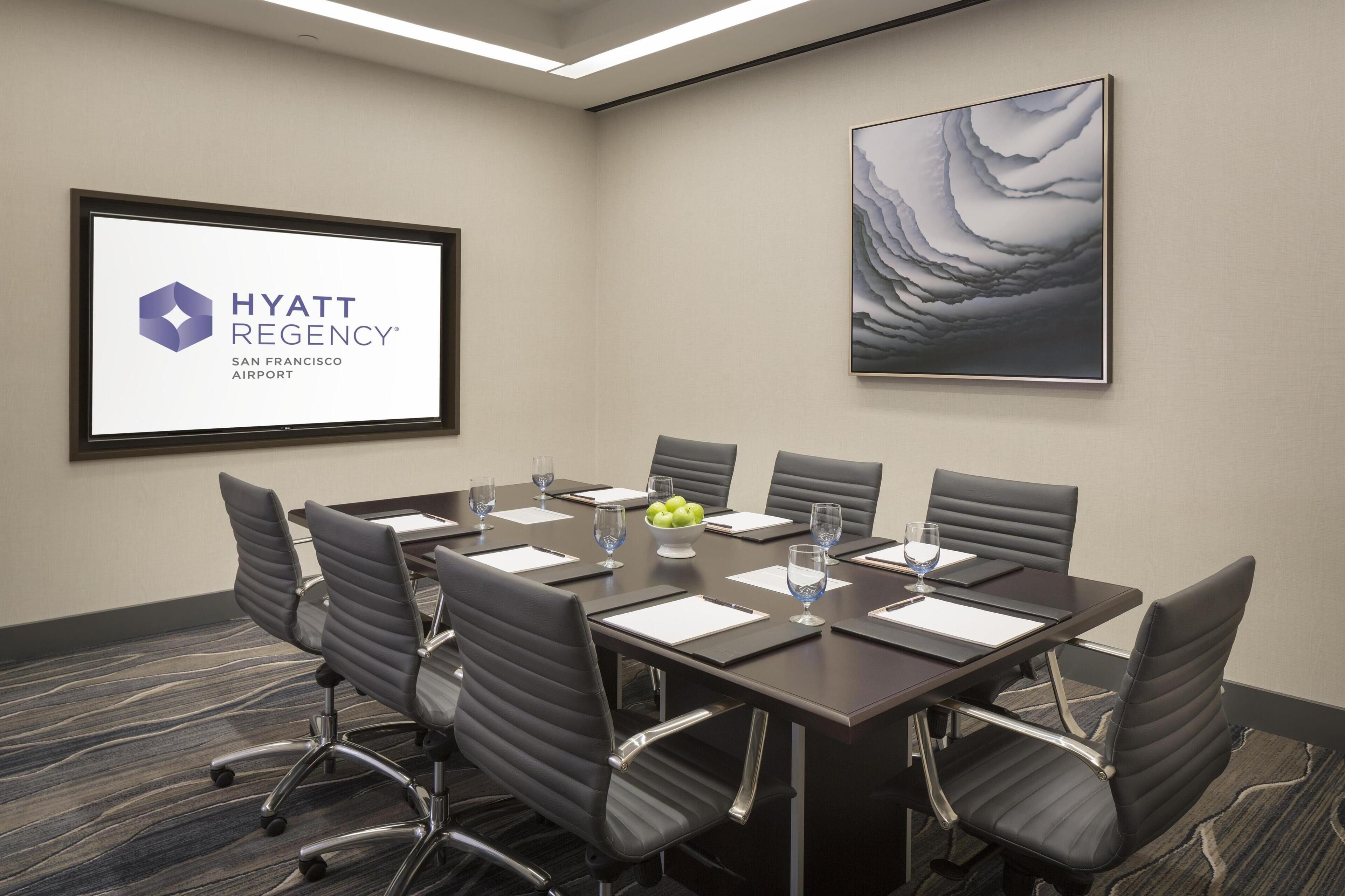 A conference room with a large screen displaying the Hyatt Regency San Francisco Airport logo, a modern painting on the wall, and a table set with chairs and water glasses.