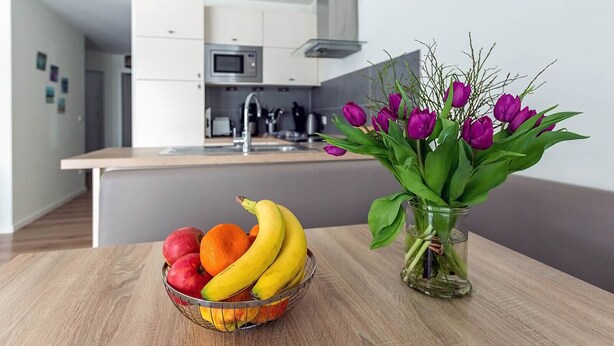 A modern kitchen with white cabinets, a microwave, and a sink. A fruit bowl with apples, oranges, and bananas is on a wooden table, and a vase with purple tulips is also visible. - Vacation apartment Ritzhagen in Willingen (Willingen)