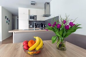 Ett modernt kök med vita skåp, en mikrovågsugn och en diskho. En fruktskål med äpplen, apelsiner och bananer står på ett träbord, och en vas med lila tulpaner syns också.