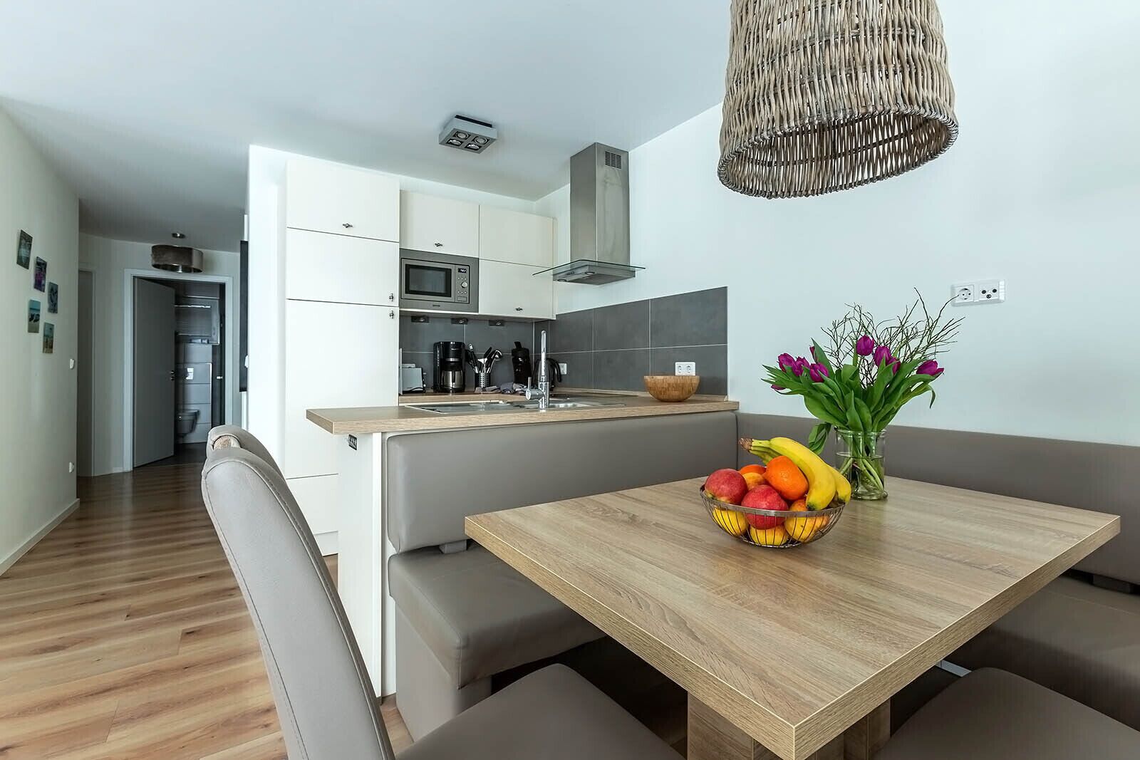 Eine moderne Küche mit weißen Schränken, einer Mikrowelle und einem eingebauten Herd. Es gibt einen Holztisch mit Obstschale und Blumenvase.