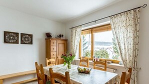 Un comedor con una mesa dispuesta para cuatro personas, rodeada de sillas de madera. Hay un armario contra la pared y una ventana con cortinas florales.