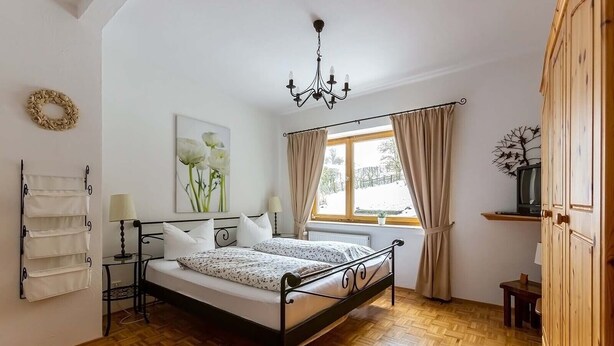 Un dormitorio con cama, mesitas de noche, candelabro, ventana con cortinas, radiador y armario de madera.