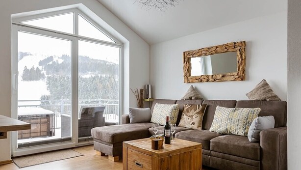 Een moderne woonkamer met een groot raam, een bruine leren bank, een houten salontafel en een rustieke spiegel aan de muur.