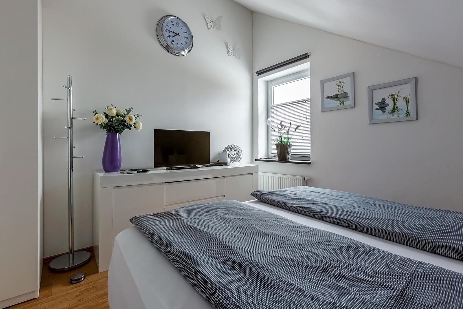Ett modernt sovrum med en säng, en tv, en vas med blommor och ett fönster med persienner.