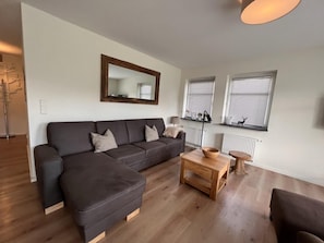 Un salon moderne avec un canapé d’angle, une table basse en bois et un grand miroir au mur.
