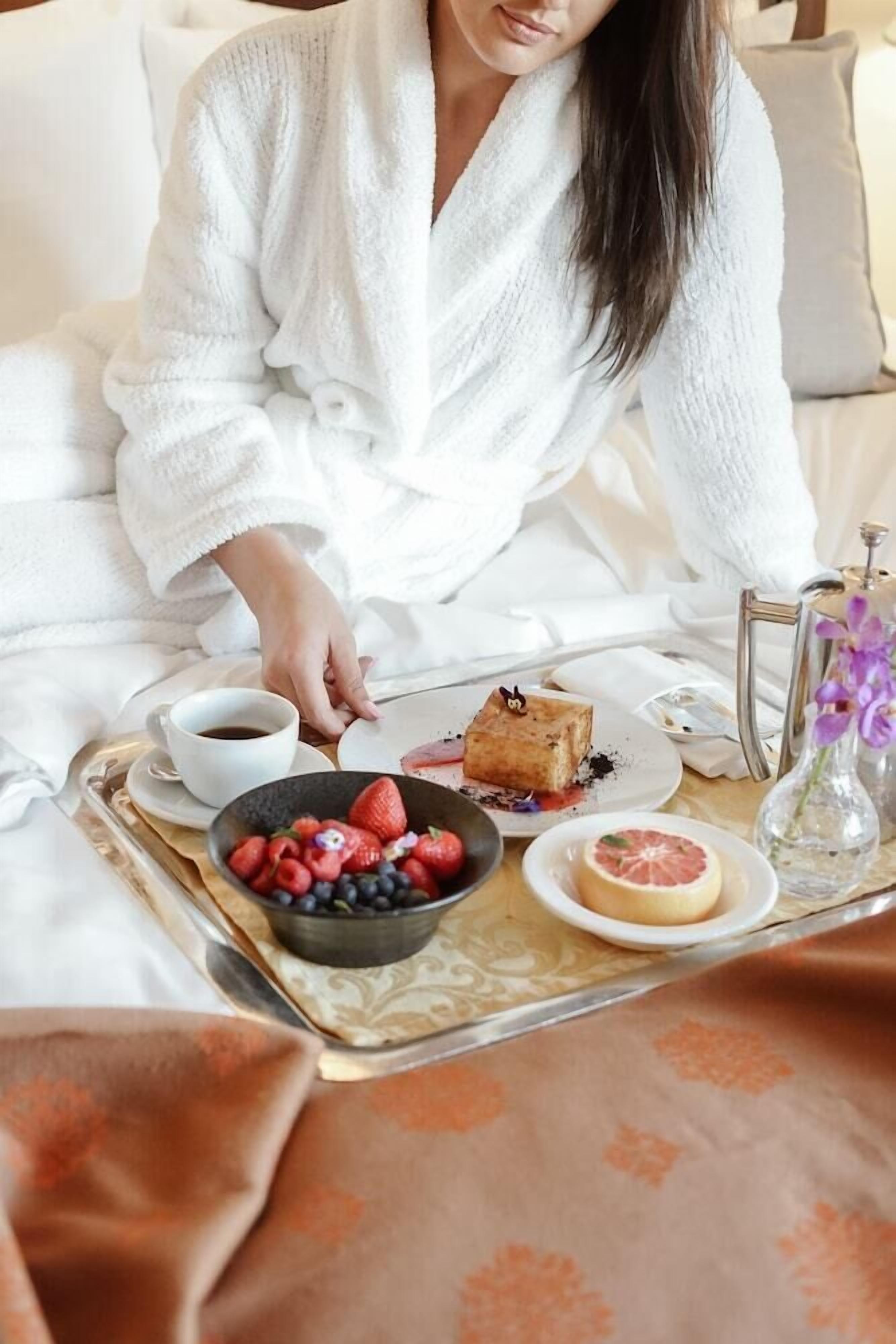 A person in a white robe sitting on a bed with a tray containing a cup of coffee, a plate with a piece of cake, a bowl of mixed berries, and a glass of water.