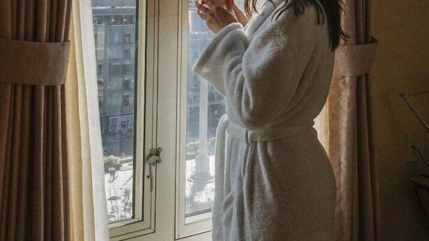 A person in a white bathrobe standing by a window with curtains, looking out at a cityscape with tall buildings.