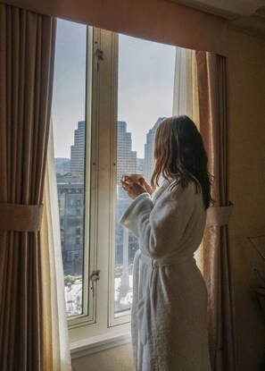 Eine Person in einem weißen Bademantel steht an einem Fenster mit Vorhängen und blickt auf eine Skyline mit hohen Gebäuden.