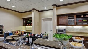 A hotel kitchen with a buffet setup, including a coffee machine, wine glasses, and a variety of food items. - 2 Connecting Suites at a  Hotel (Houston)
