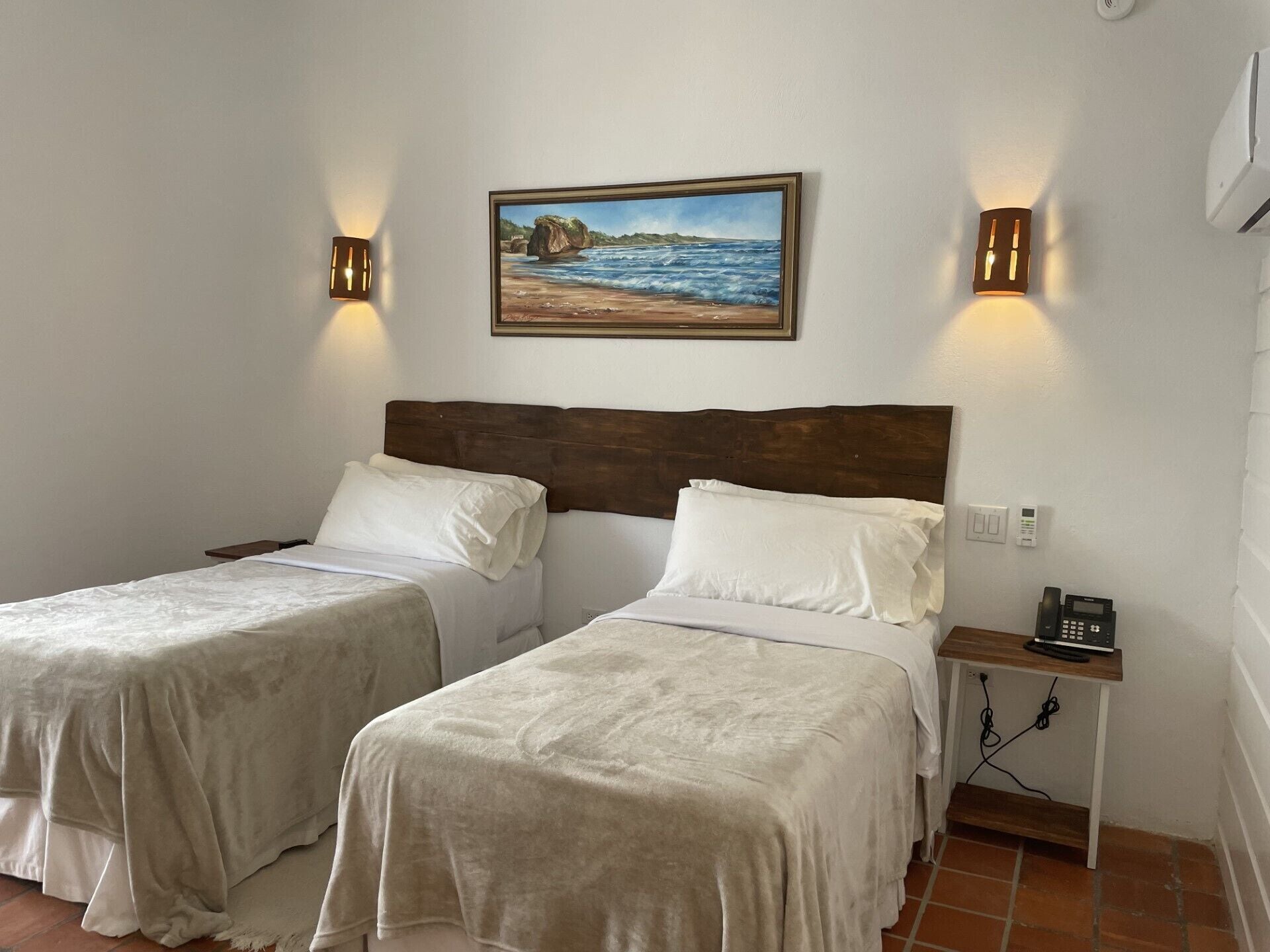 Une chambre double avec deux lits, un tableau représentant une scène de plage, des lampes fixées au mur, une table de chevet avec un téléphone et une prise de courant.