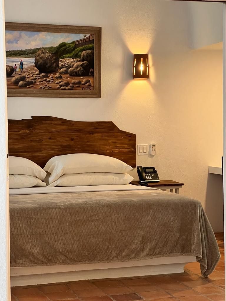 Une chambre d’hôtel avec une tête de lit en bois, un lit avec du linge de lit blanc, un tableau au mur, une table de chevet avec un téléphone et un luminaire fixé au mur.