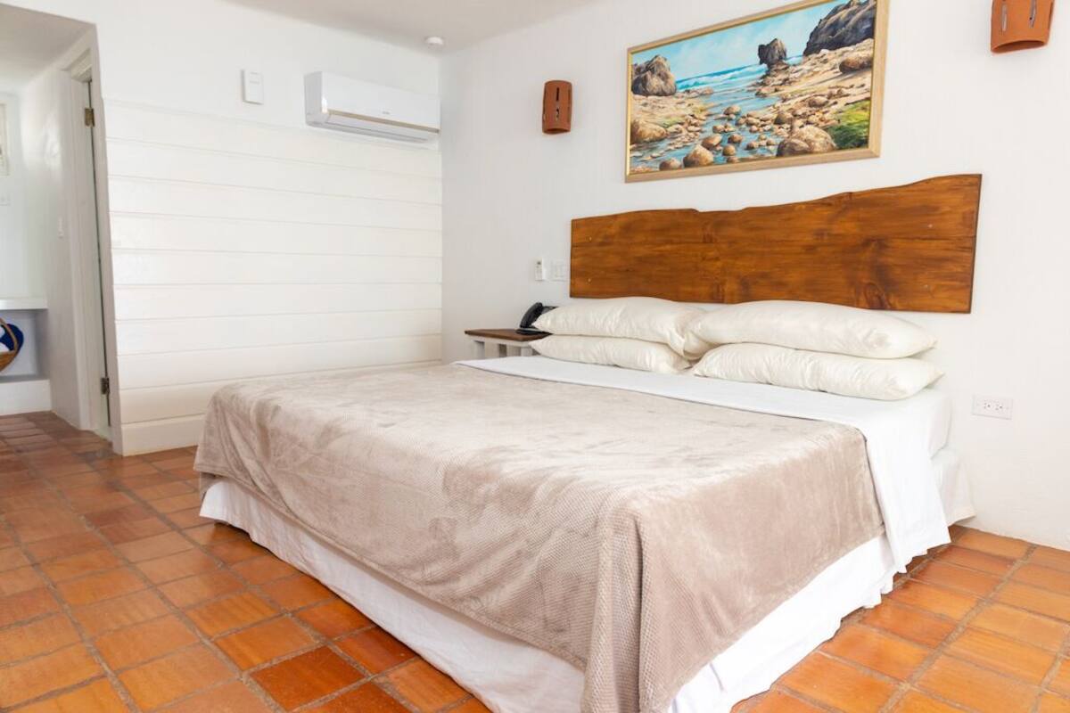Ein Schlafzimmer mit einem großen Bett, weißen Wänden, einem hölzernen Kopfteil, einem Strandbild und einem Fliesenboden.