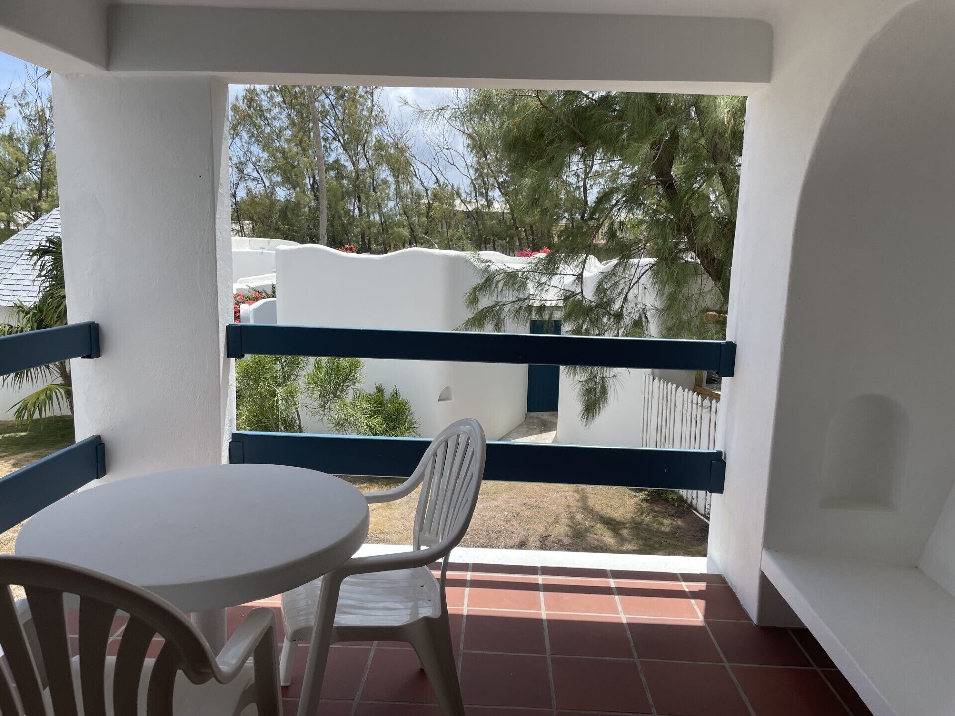 A balcony with a white table and chairs, a view of a white building and trees, and a tiled floor.