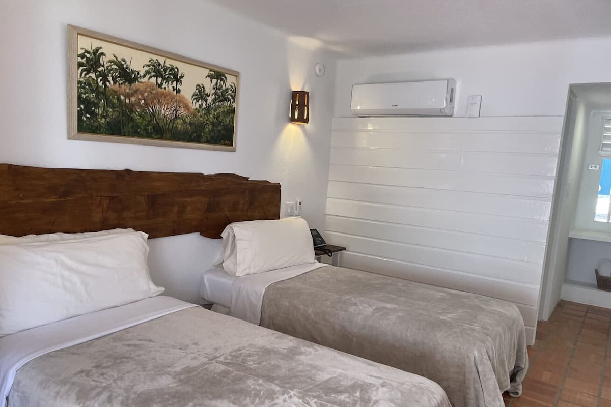 Ein Hotelzimmer mit zwei Betten, einem Holzkopfstück, einem Bild mit tropischer Szenerie, einer Klimaanlage und einem gefliesten Boden.