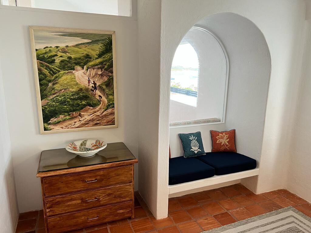Una habitación con un aparador de madera, un cuadro de un paisaje, un banco con cojines y vistas a través de un ventanal arqueado.
