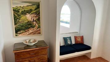 Ein Zimmer mit einem Holzkoffer, einem Landschaftsbild, einer Bank mit Kissen und Blick durch ein Rundbogenfenster.