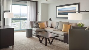 A modern hotel room with a sectional sofa, a dark coffee table, a desk, and a large window offering a view of the city.