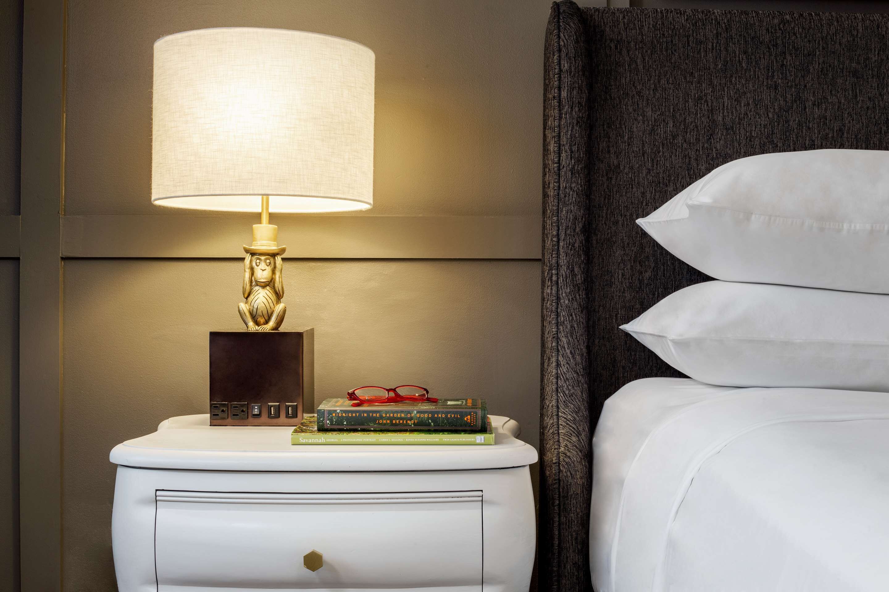 Un lit bien fait, avec du linge de lit blanc, une lampe de chevet à la base dorée et une pile de livres sur une table de chevet.