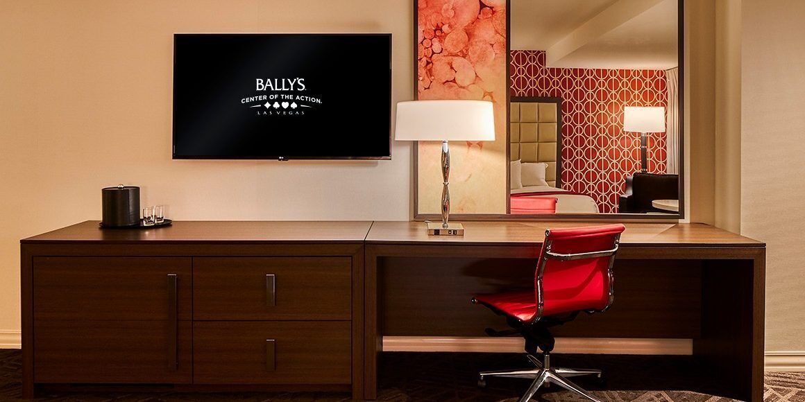 A hotel room with a wooden desk, a red chair, a flat-screen TV displaying "BALLY'S CENTER OF THE ACTION LAS VEGAS", a large mirror, and a patterned wall.