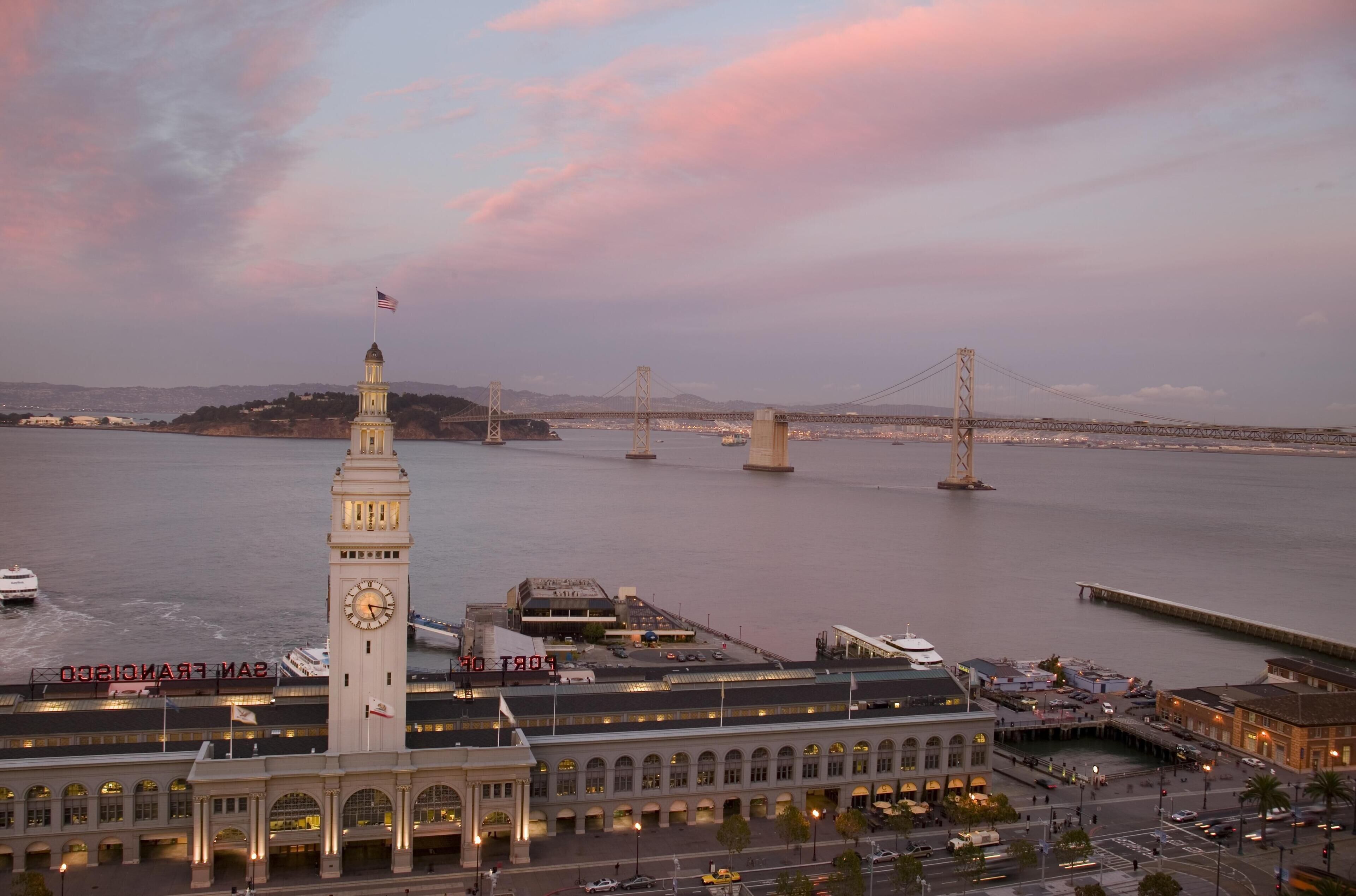 A waterfront building with a clock tower, a large bridge, and a body of water.
