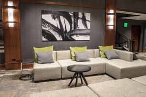 Un hall d’hôtel moderne avec un canapé d’angle, une petite table ronde et un grand tableau abstrait accroché au mur.
