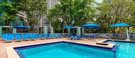 Una piscina con sillas y sombrillas azules, rodeada de árboles y edificios modernos.