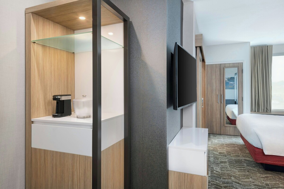 Habitación de hotel con cama, televisor montado en la pared, cafetera y repisa de vidrio.