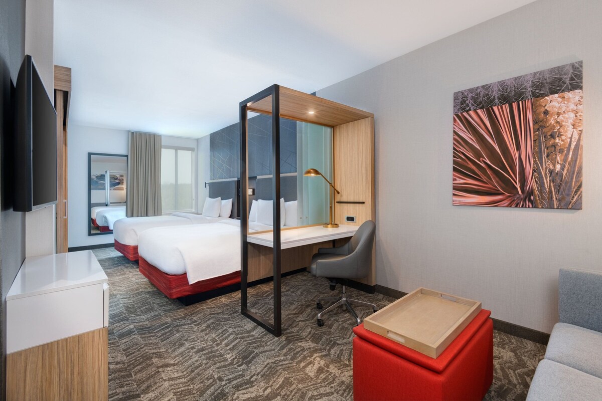 Una habitación de hotel con cama, escritorio, silla, televisor, obras de arte y un reposapiés rojo.