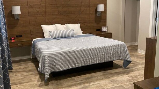 A hotel room with a bed, wooden headboard, bedside lamps, a nightstand, a wooden floor, and a door.