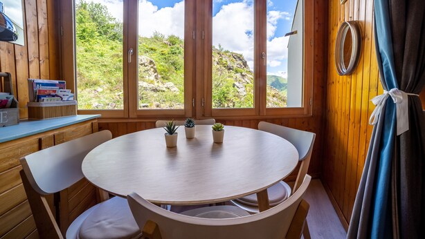 A wooden cabin with a round dining table, four chairs, and a view of a mountainous landscape through large windows. - L'Eclose - Studio au pied des pistes - Alpe d'Huez (Huez)