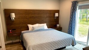 A hotel room with a large bed, wooden headboard, bedside lamps, a nightstand, and a view of the outdoors through a window.