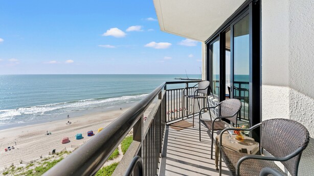 Un balcon avec vue sur la plage et l’océan, des fauteuils en osier et une table agrémentée d’un vase.