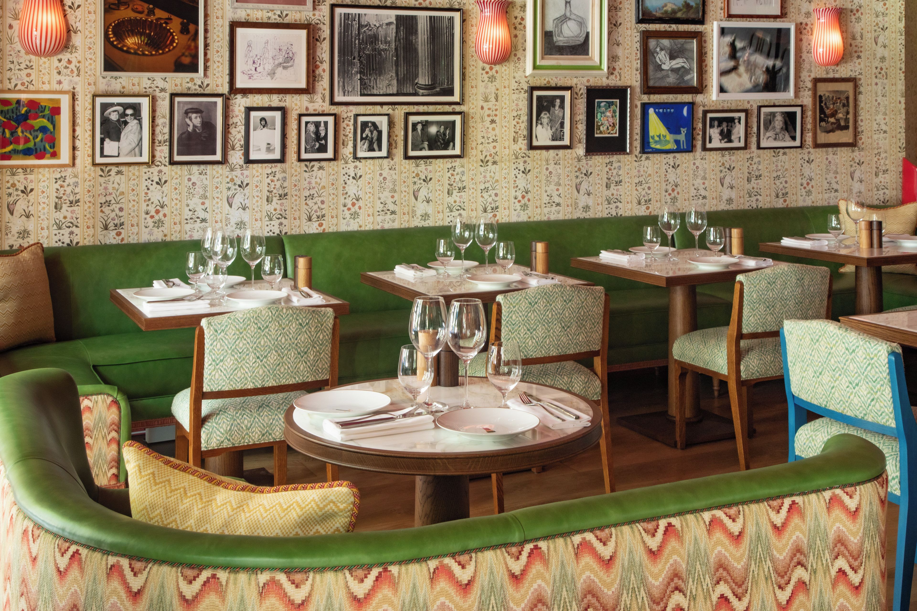 Interior de restaurante com assentos estofados em verde, mesas postas com copos de vinho e pratos, e uma parede decorada com obras de arte emolduradas.