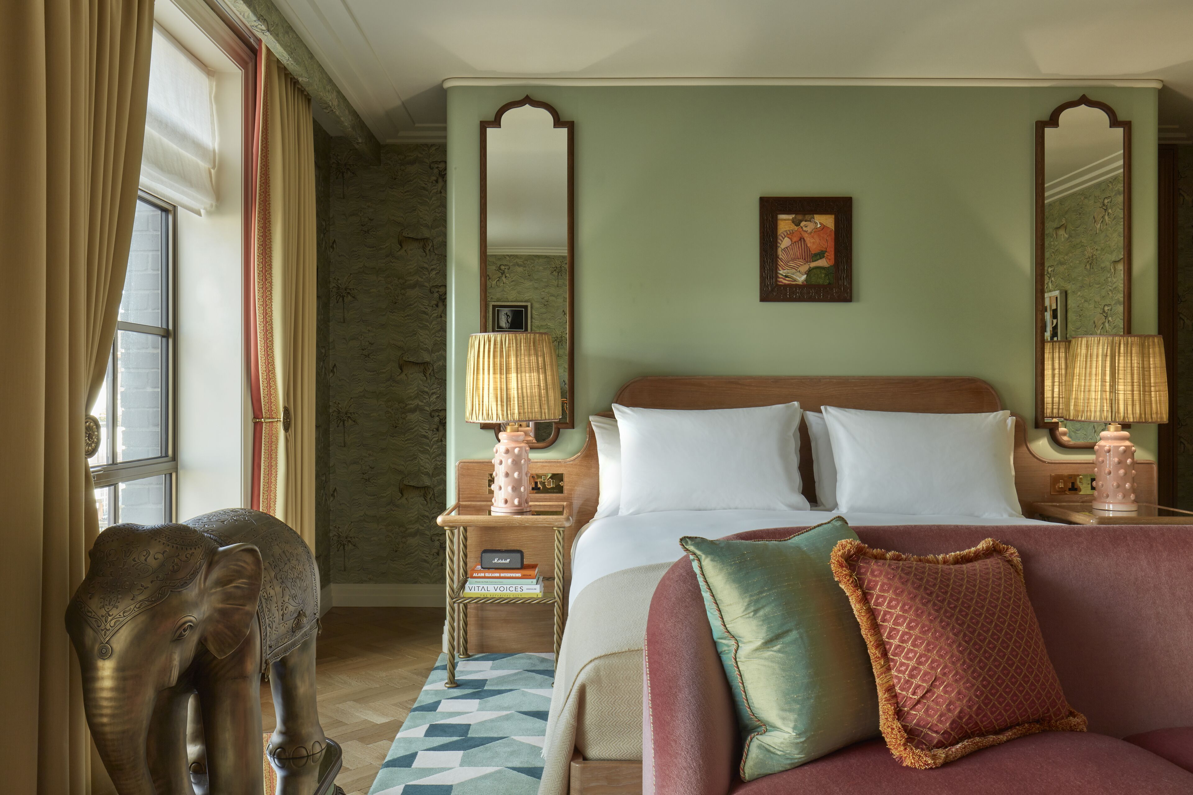 A hotel room with a large bed, two bedside lamps, a nightstand with books, a decorative elephant statue, and a patterned rug.