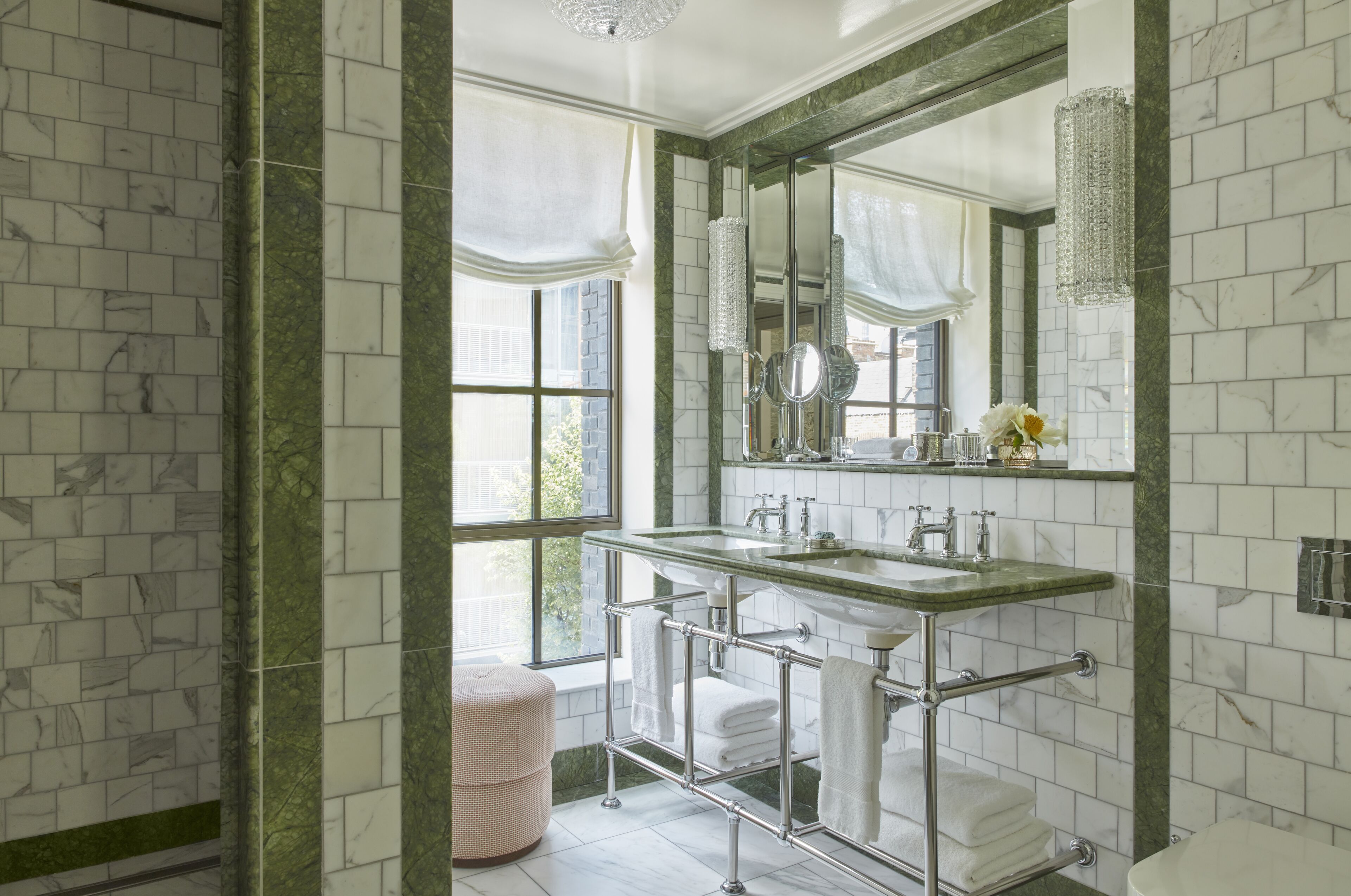 A bathroom with a marble-tiled wall, a large mirror, a double sink, and a towel rack.