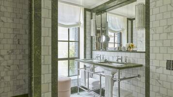 A bathroom with a marble-tiled wall, a large mirror, a double sink, and a towel rack.