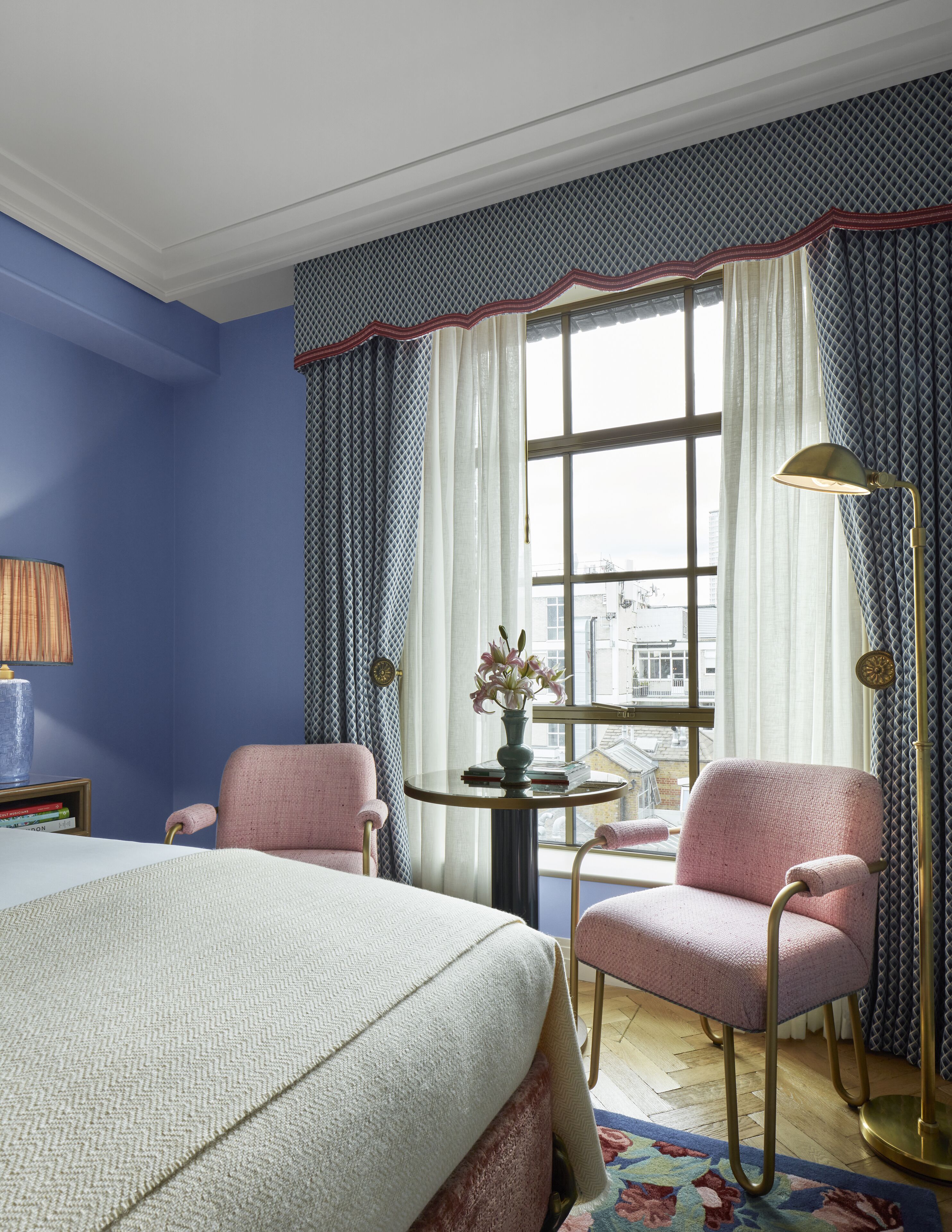 A hotel room with a large bed, two pink armchairs, a small round table with a vase of flowers, and a window with curtains.