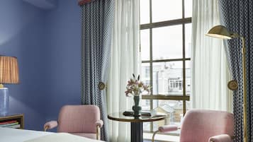 A hotel room with a large bed, two pink armchairs, a small round table with a vase of flowers, and a window with curtains.