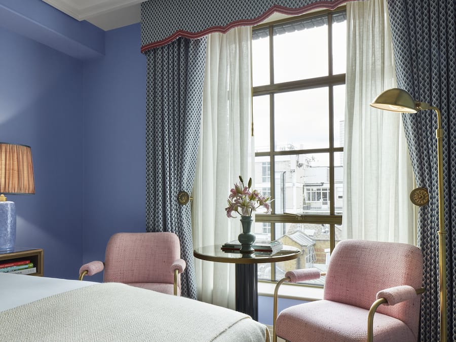 A hotel room with a large bed, two pink armchairs, a small round table with a vase of flowers, and a window with curtains.