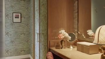 Ein Zimmer mit einem hölzernen Schminktisch, einem Spiegel, einer Vase mit Blumen und einem gemusterten Sessel.