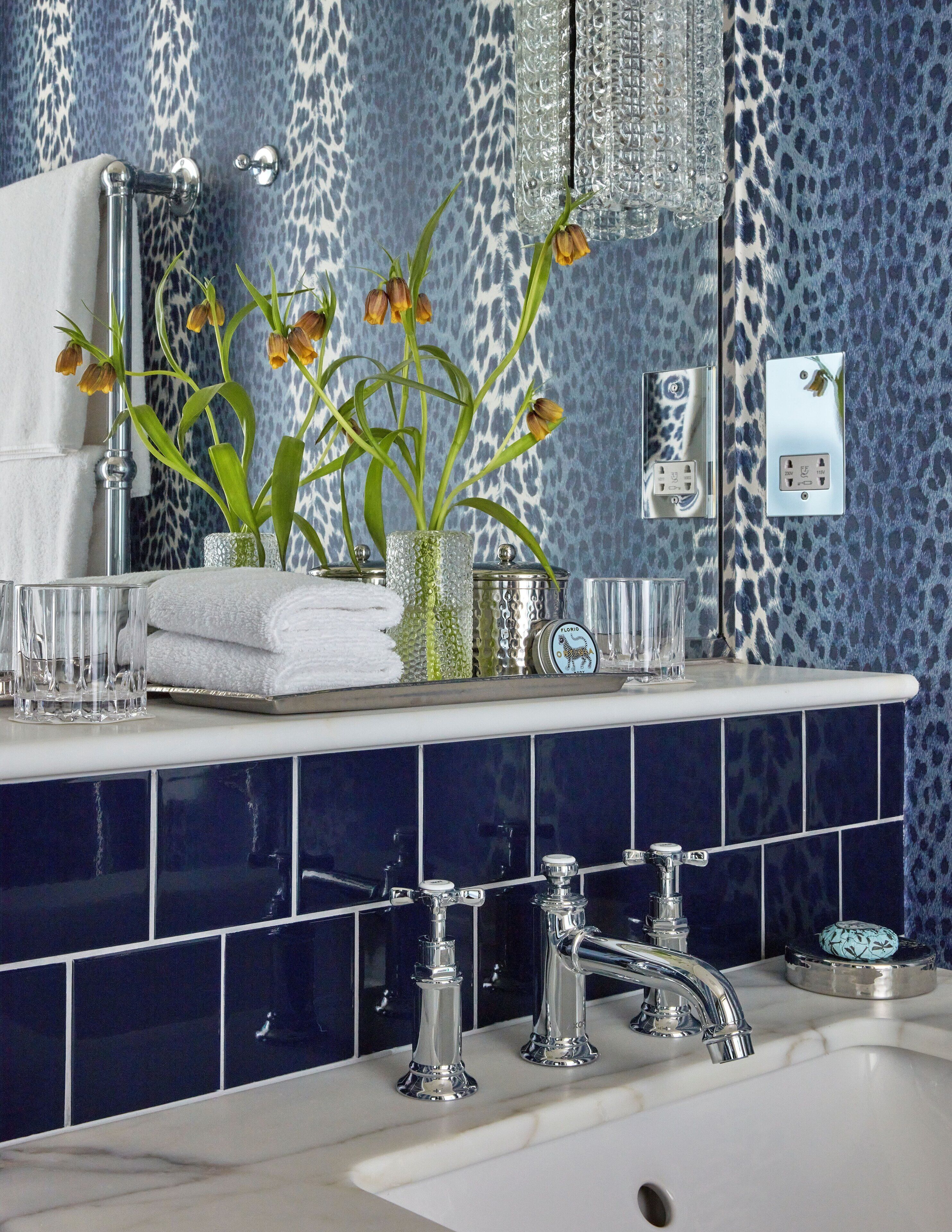 A bathroom with a blue patterned wall, a white sink, two chrome faucets, and a mirror above the sink.