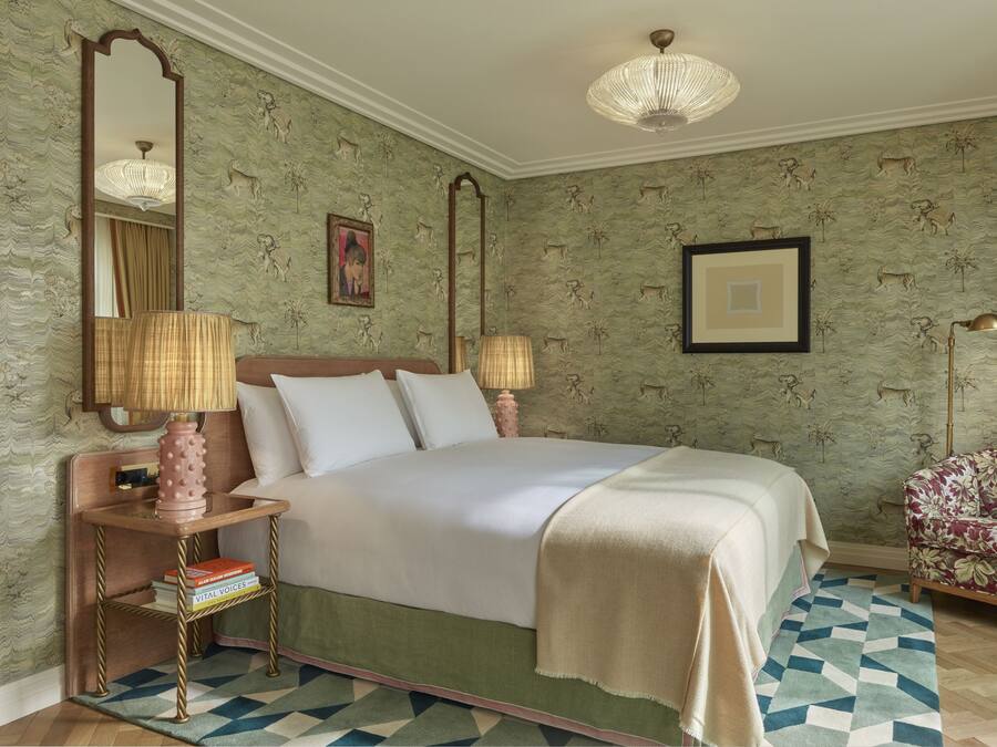 A neatly arranged bedroom with a bed, bedside lamps, a nightstand with books, a patterned chair, and a large mirror.