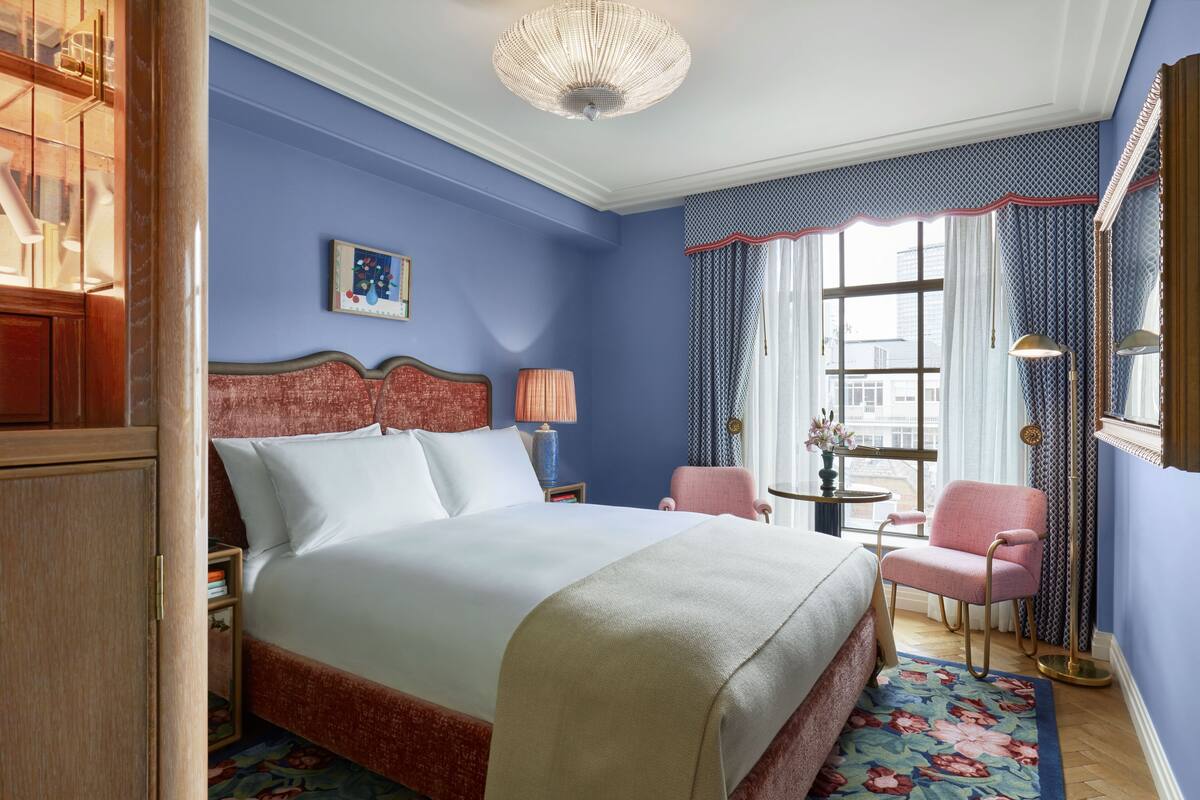 Una habitación de hotel con una cama grande, un armario de madera, una mesita con lámpara, una silla rosa y una ventana con cortinas.
