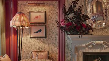 A room with a fireplace, a lamp, a chair, an ottoman, and framed artwork on the wall.