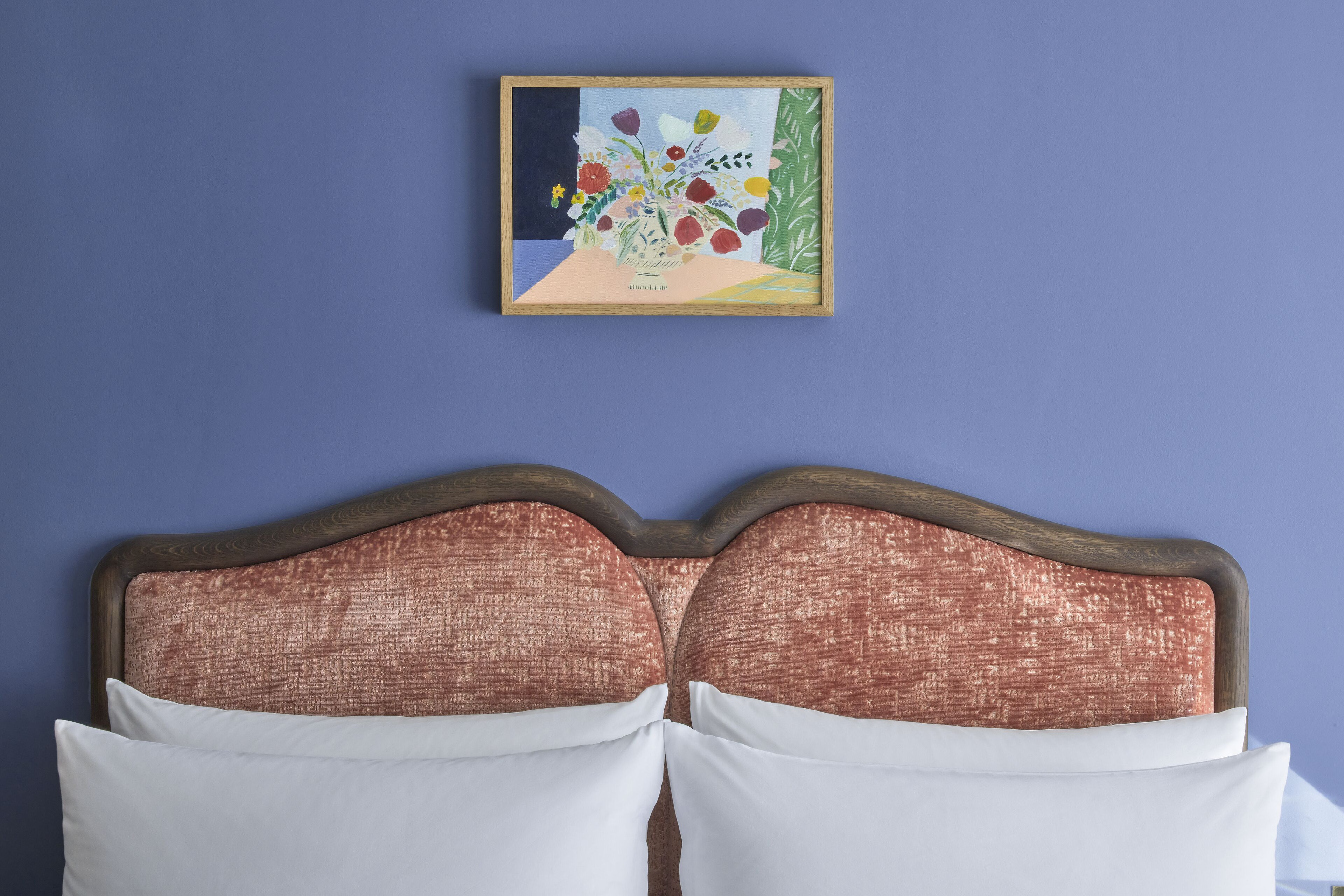 Ein Bett mit weißen Kissen und einem Holz-Kopfstück vor einer blauen Wand. Über dem Bett hängt ein gerahmtes Bild mit Blumen.