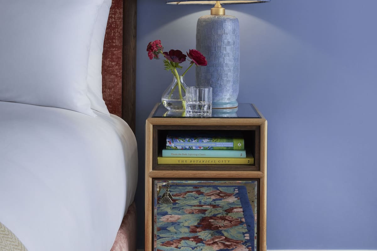 Una cama bien hecha con ropa blanca, una mesita de noche de madera con lámpara, un florero con flores e impresión botánica enmarco.
